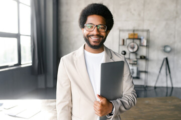 A Brazilian businessman smiles warmly, holding a laptop in one hand, standing in a chic office environment that reflects a modern, agile business culture. A blend of friendliness and competence