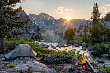 Camping tent set up by a river in a mountain valley with a campfire and the sun setting behind the peaks.