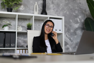 A poised Asian businesswoman in the corporate office manages tasks on her computer and smartphone,...