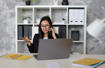 A young Asian businesswoman works joyfully in her office, utilizing her laptop for online tasks and communication, displaying professionalism and positivity.