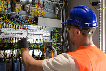 an electrician in a blue helmet, yellow glasses and an orange vest measures electric current with a...