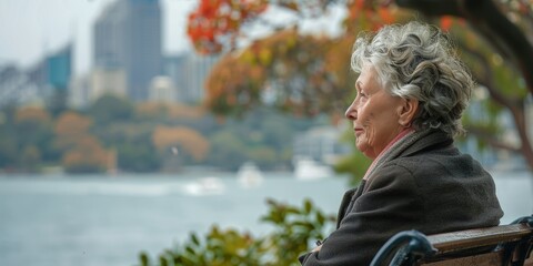 An elderly woman sits on a bench on the shore of the bay and looks into the distance.