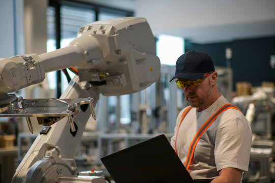 engineer in white t-shirt programming robot on assembly line using laptop blurred background