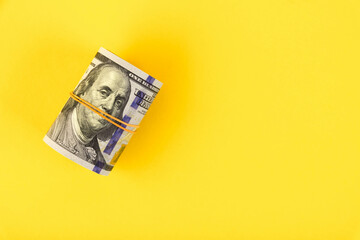 A roll of hundred-dollar American bills is tied with elastic band on a yellow background. Copy space