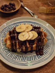 Waffle with melted hot chocolate and banana accompanied by peanuts with cocoa on a wooden table