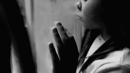 One unhappy somber little girl feeling depressed during hard times. Close-up child's face and hand leaning on glass window feeling lonely and solitude in black and white, monochromatic