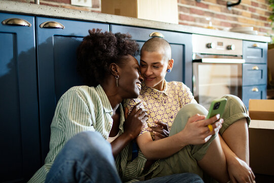 Lesbian couple smiling and embracing in a cozy kitchen with one person holding a smartphone