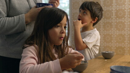 Pensive little girl eating yogurt from bowl in kitchen with mother and little brother in the...