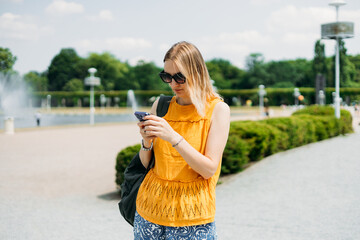Happy cheerful young blond woman with backpack walking on city street checks her smartphone....