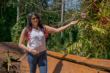Woman walking in the garden of the open-air museum in Minas Gerais, with many trees and vegetation around, a large tree trunk worked roughly and holding a beautiful flower.