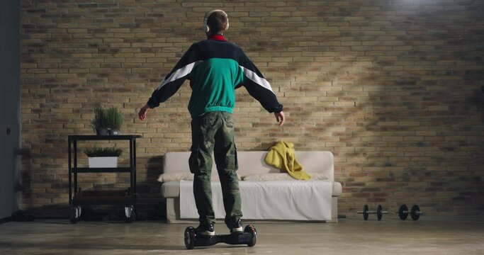 Young teenager wearing headphones is having fun to listen his favorite music playlist and feeling free with open arm while rides technological gadget hoverboard at home.