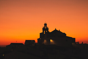 Iglesia católica de Nuestra Señora de las Flores en Sanlúcar de Guadiana, Huelva, España. Silueta del campanario de la iglesia en un cielo anaranjado durante la puesta de sol. - obrazy, fototapety, plakaty