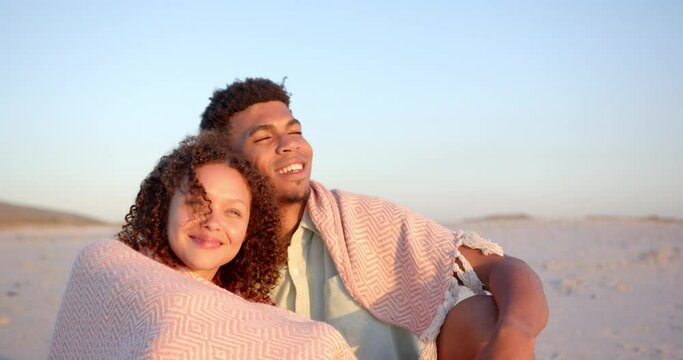 Biracial couple wrapped in a blanket, enjoying a beach at sunset