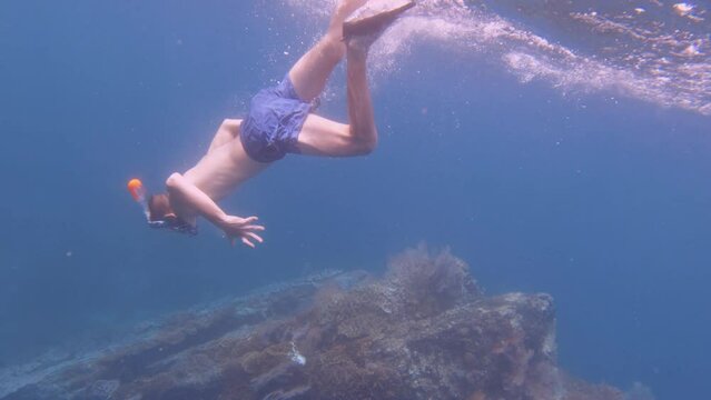 a noob newbie white Caucasian teen male boy wearing snorkel mask and fin duck free diving kicking down into the deep blue ocean water to beautiful pink red gorgonian sea fan and finger leather corals