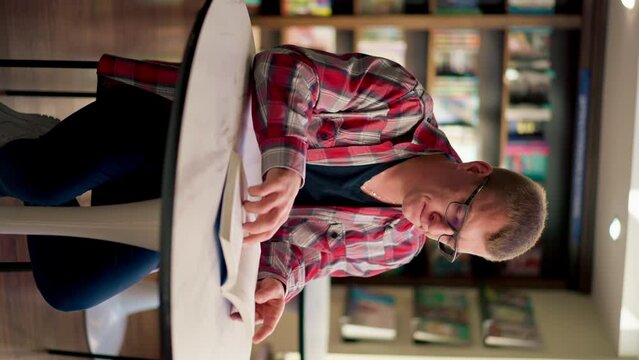 vertical video male professor in glasses sits in a scientific and technical library reads a book and prepares write a dissertation