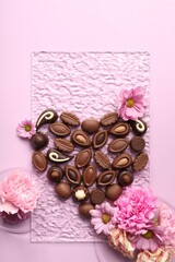 Heart made with delicious chocolate candies and beautiful flowers on pink background, flat lay