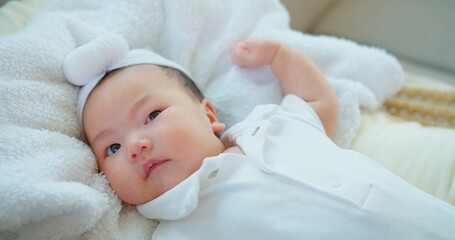 A contented baby with sparkling eyes and a soft headband lies on a plush fluffy white blanket,...