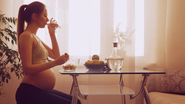 young pregnant woman sitting at the table eats fruit salad and drinks water  