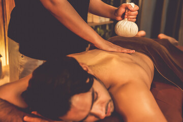 Hot herbal ball spa massage body treatment, masseur gently compresses herb bag on man body. Tranquil and serenity of aromatherapy recreation in warm lighting of candles at spa salon. Quiescent - obrazy, fototapety, plakaty