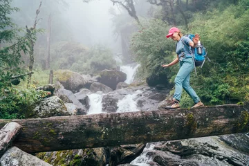 Photo sur Plexiglas Makalu Young woman with backpack and trekking poles crossing wooden bridge near power mountain river waterfall during Makalu Barun National Park trek in Nepal. Mountain hiking and active people concept image