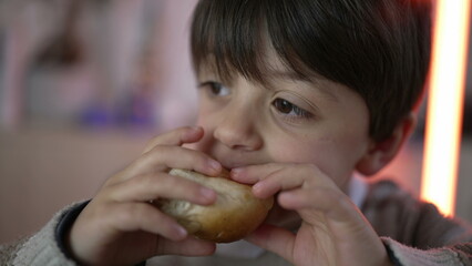 Close-up child's hand holding a piece of bread and taking a bite of rich carb food. Little boy snacking