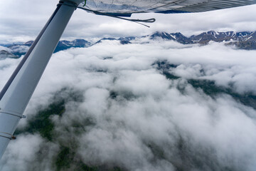 Point of View (POV) from under the wing of a Cessna fixed wing airplane with seaplane wing rope. Flying over a cloud filled sky in Alaskan wilderness. Bush pilots are primary way of accessing Alaska. - obrazy, fototapety, plakaty