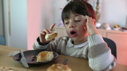 Child eating piece of bread seated at table. 5 year old boy snacking rich carb food