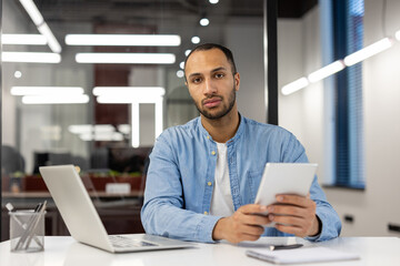 Fototapeta na wymiar Portrait of a young serious Muslim man sitting in the office at a desk with a notebook and documents, holding a tablet in his hands and looking seriously into the camera