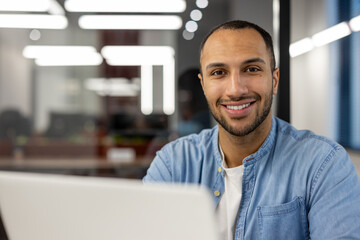 Close-up portrait of young smiling male freelancer and student working and studying in modern office, sitting at laptop and looking at camera - obrazy, fototapety, plakaty