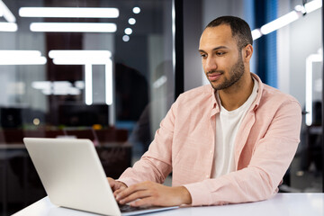 A young hispanic man is sitting in a modern office at a desk in a pink shirt, working and studying on a laptop