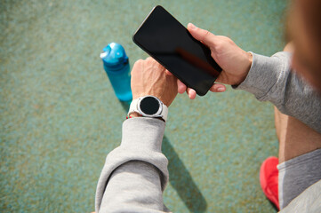 Male athlete using smartphone with black mockup screen, checking mobile app on his wrist watch, after workout outdoor