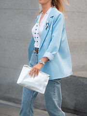 A woman in a light blue blazer and jeans with a silver clutch in her hand, wearing an elegant white...