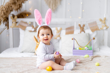 cute happy baby girl in bunny ears on the bed at home with a rabbit and painted eggs, easter...