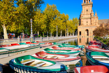 Obraz premium Green and red boats for hire sit in the water ready for visitors in the morning at the Plaza de Espana public square and park in the Andalusian city of Seville, Spain.