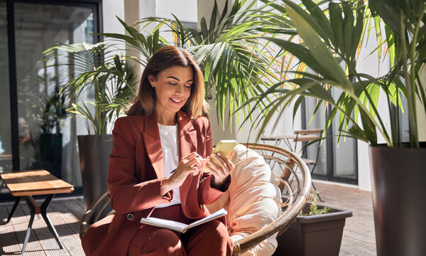Happy smiling elegant mature middle aged business woman wearing suit holding cellphone using mobile cell phone looking at smartphone sitting in comfortable chair in sunny office with green plants. - Powered by Adobe