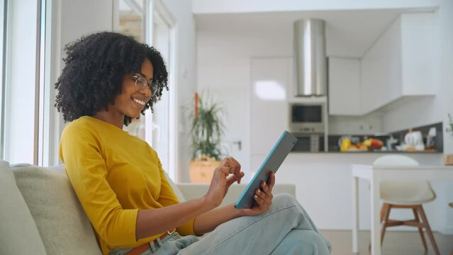 Happy young African American woman sitting on couch at home using digital tablet. Smiling lady relaxing on sofa looking at tab technology device holding pad computer in hands in living room.