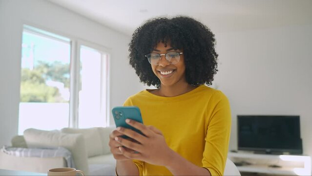 Happy smiling young African American woman customer sitting at home table looking at cellphone while holding mobile cell phone in hands texting using smartphone scrolling media, shopping online.