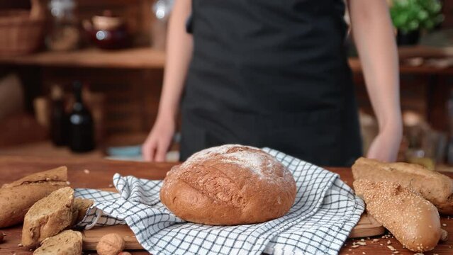 Professional baker in black apron putting fresh baked bread on wooden table, close-up view in bakery