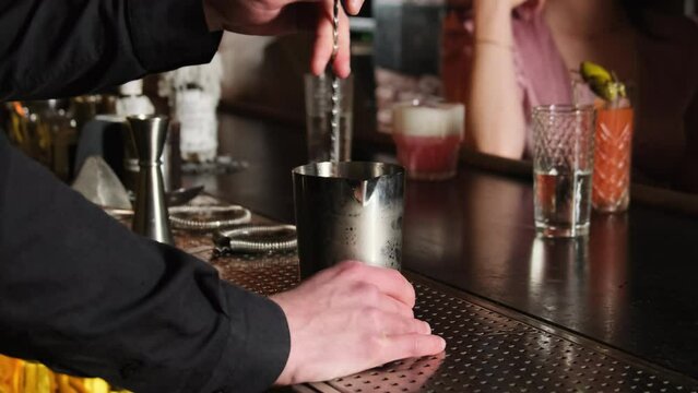 Skillful bartender holds long bar spoon and mixes drink in cocktail glass. Guests waiting for new tasty beverages in background