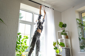 Woman hangs transparent tulle curtains on large windows in the house inside the interior. Spring...