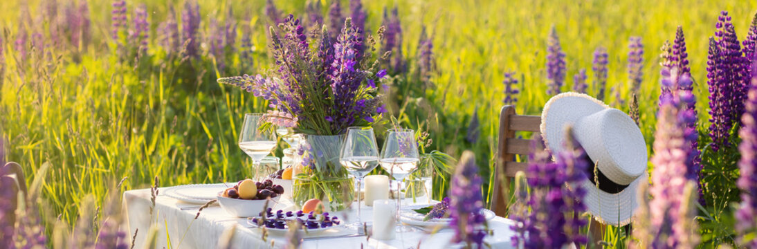 Beautiful romantic outdoor wedding decor in field. Table decorated with purple lupines flowers. Wineglasses with white wine. Sunset, summer, golden hour. Perfect surprise date for loving couple banner
