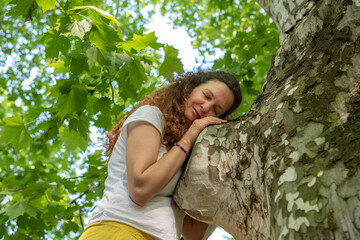 Beautiful woman with gorgeous curly hair hugging tree, dressed in white and yellow combination, hugging plane tree, eyes closed. Concept: active, healthy life, in love with nature
