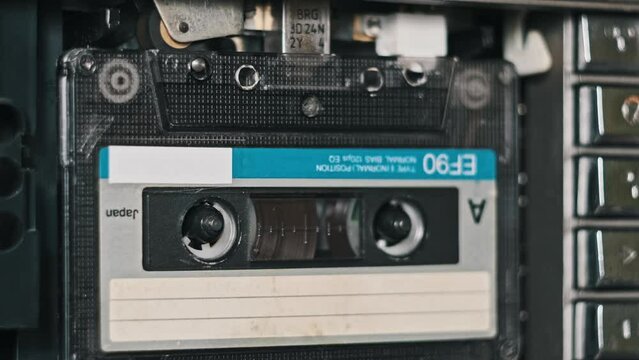 An audio cassette in a tape recorder is played in close-up. Playing audio cassette. Vintage record sound in a retro player. Recording or playback conversations. Retro tape reels rotate in deck. 80s