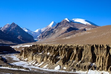 Experience the stunning contrast of a brown land formation against snow-capped mountains and a...