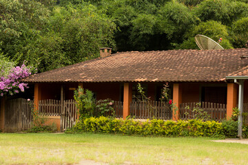 Fototapeta na wymiar Casa de fazenda com varanda, telhado de telha de barro cercada de vegetação. 