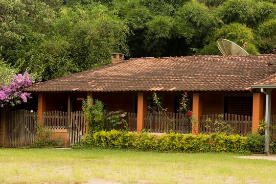 Casa de fazenda com varanda, telhado de telha de barro cercada de vegetação. 