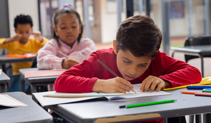 Biracial boy in red focuses intently on his drawing in a school classroom setting
