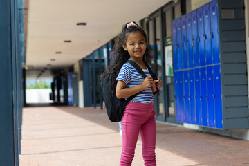 Biracial girl with a backpack stands smiling at school