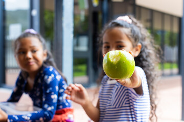 Two biracial girls are sharing a moment in school, one holding a green apple towards the camera