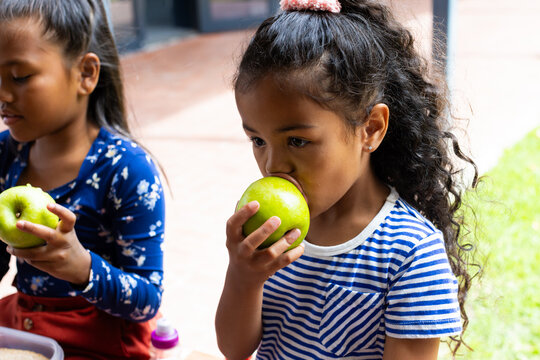 Biracial girls enjoy a healthy snack outdoors in school, one biting into a green apple